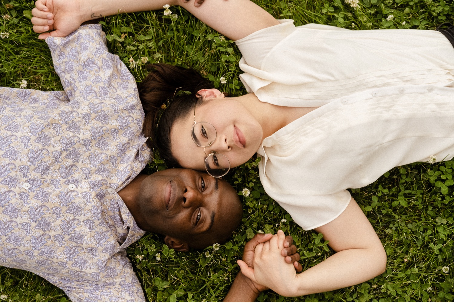 Couple laying in the grass heads next to each other holding hands