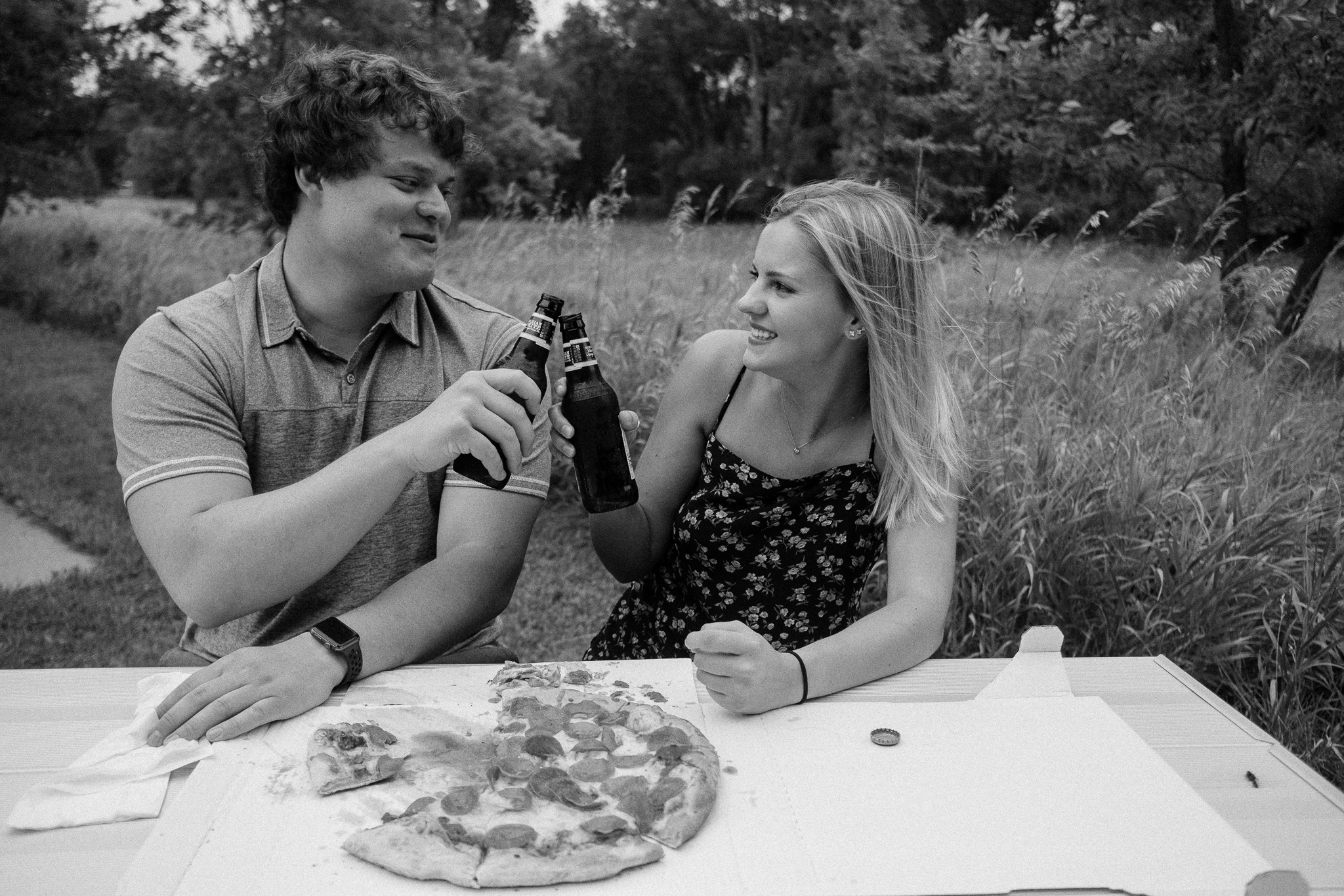 Couple clinking beer bottles at a picnic table while eating pizza.