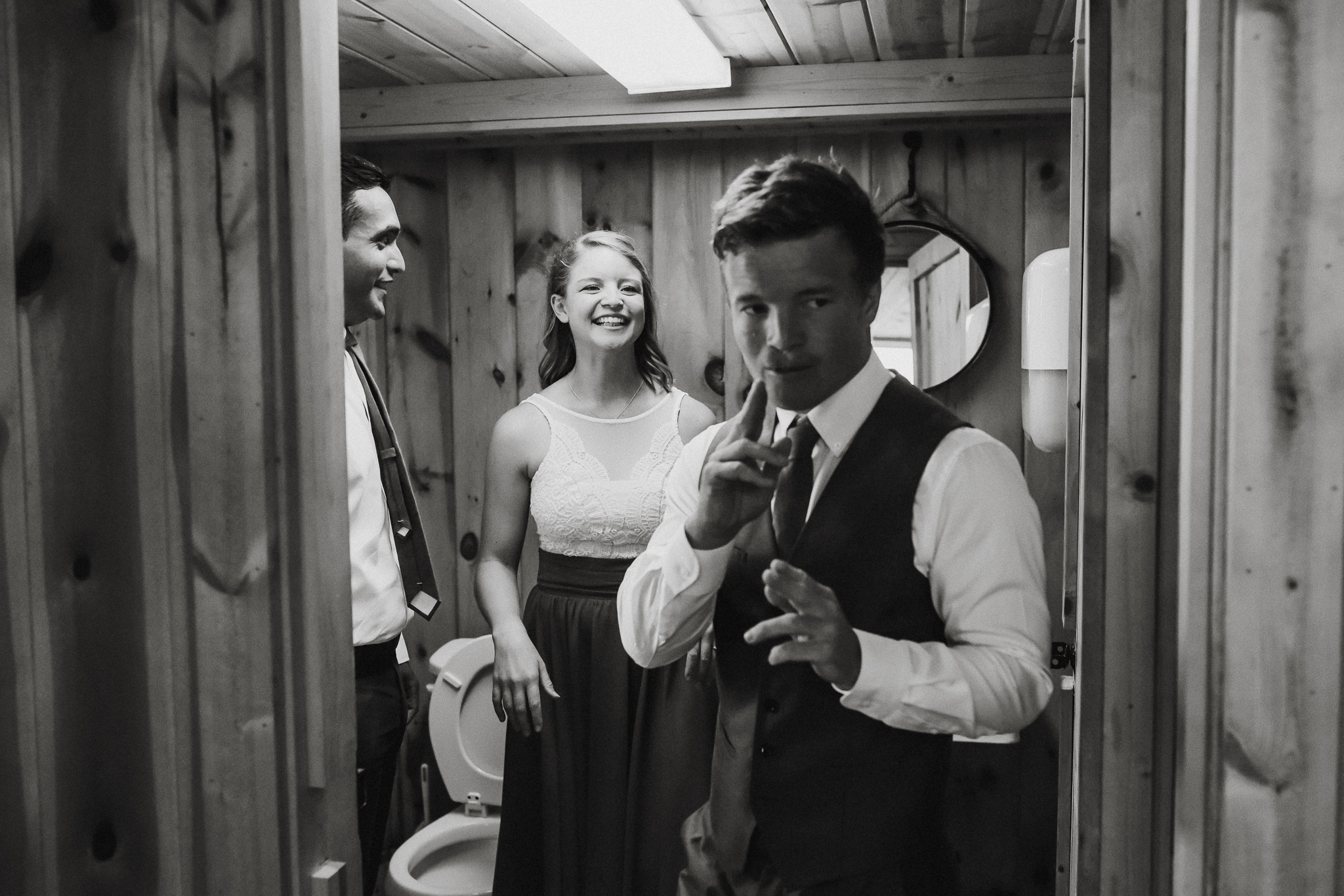 Groom exiting bathroom shooting finger guns. Groomsman and a bridesmaid behind groom laughing. Wedding in St. Charles MN.