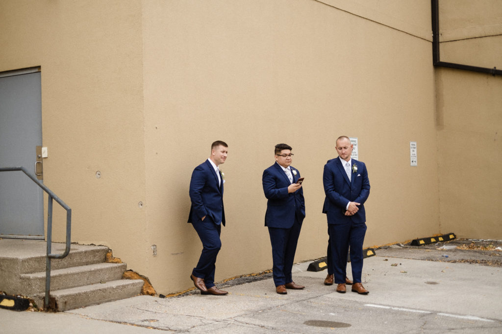 Groomsmen standing by a wall in a Des Moines Iowa alley waiting for the groom to be done with portraits. 