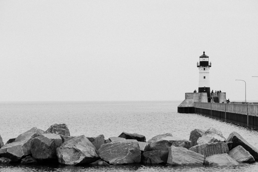 Black and white photo of rocky shoreline with lighthouse in the background. Canal Park, Duluth MN