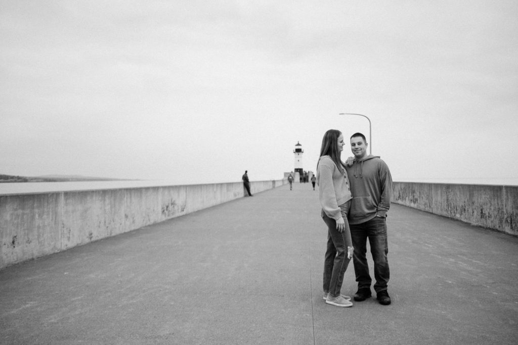 Black and white photo of couple standing on walkway with the lighthouse in the background. Duluth MN