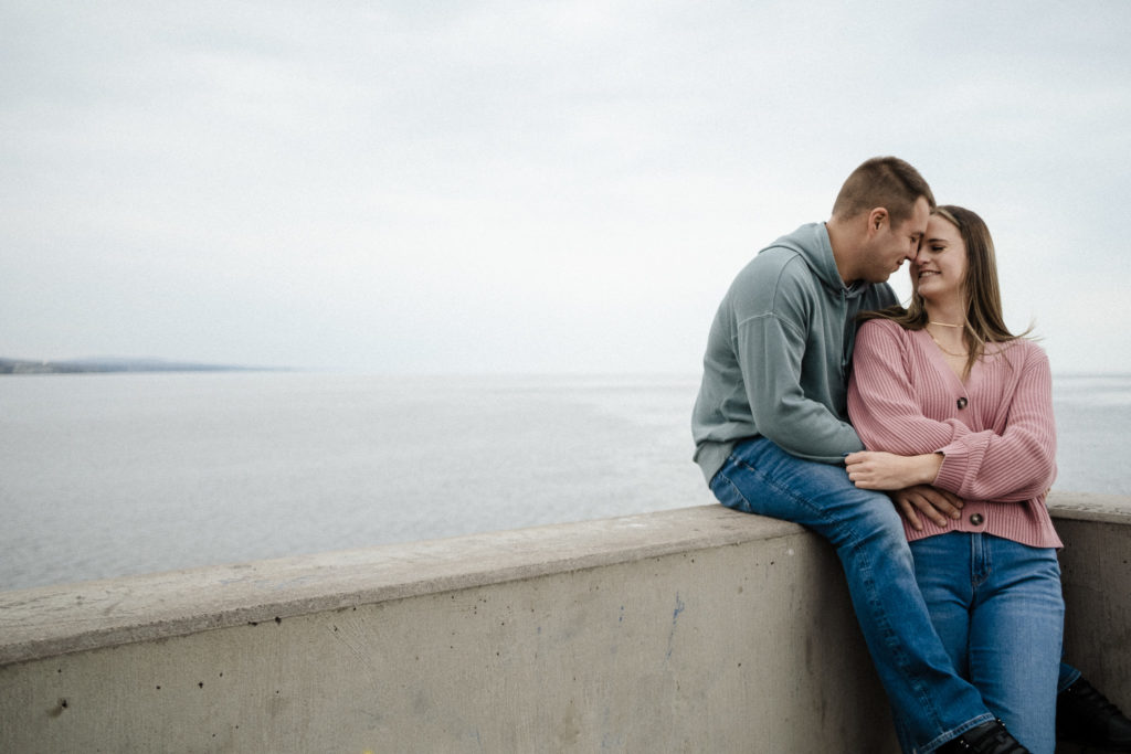 Couple sitting on the ledge by the lakeside on a cloudy day, foreheads touching and smiling at each other.