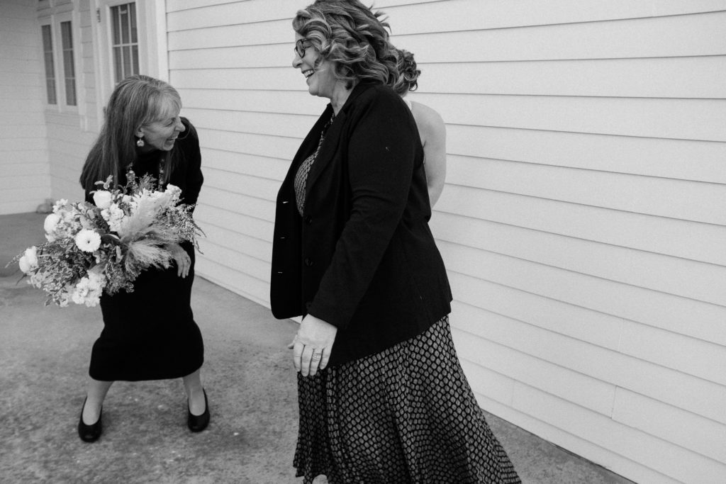 Black and white image of the bride and her mom laughing, while the bride's aunt walks past them laughing. 
