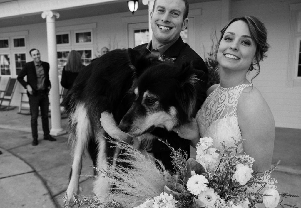 Black and white image of bride and groom holding their dog and smiling at the camera. 