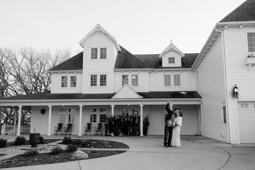 Black and white image of bride and groom kissing in the foreground while their families cheer and should from the background in front of their wedding venue, the Oakwood Inn at Okoboji.