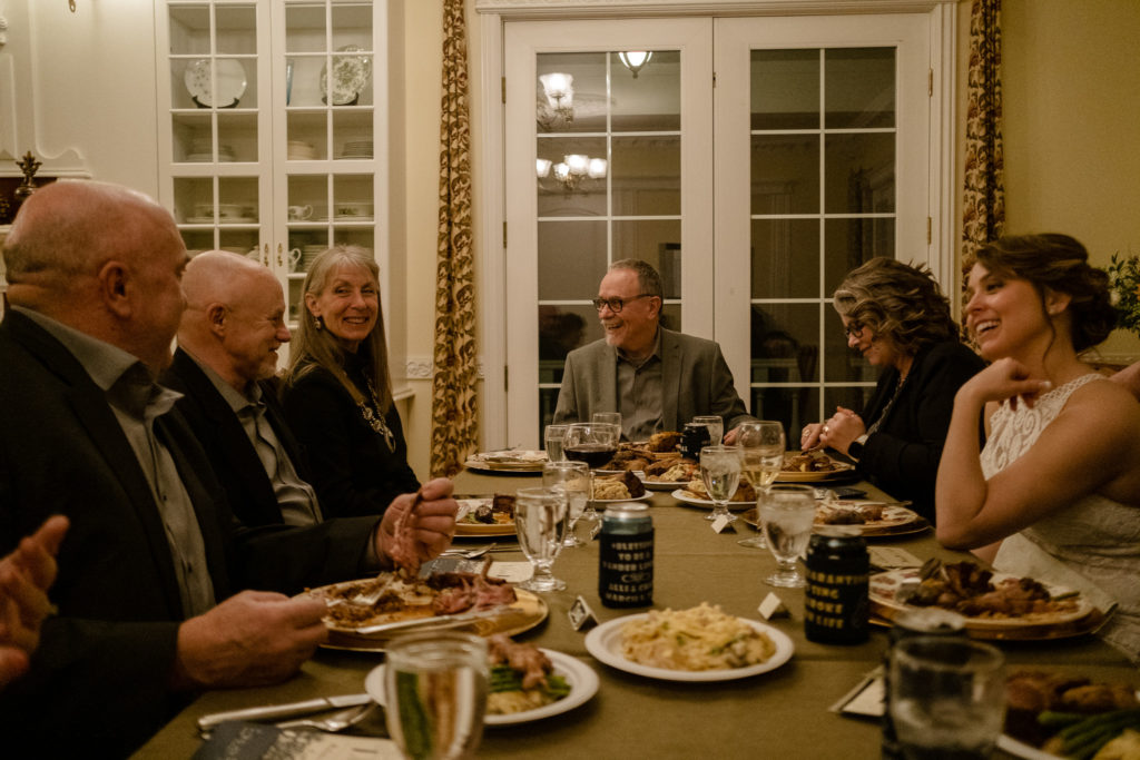 Image taken from the head of a long, family style dinner table. Frame includes the bride and family members, laughing and chatting with each other, plates full of food and drinks lining the table. 