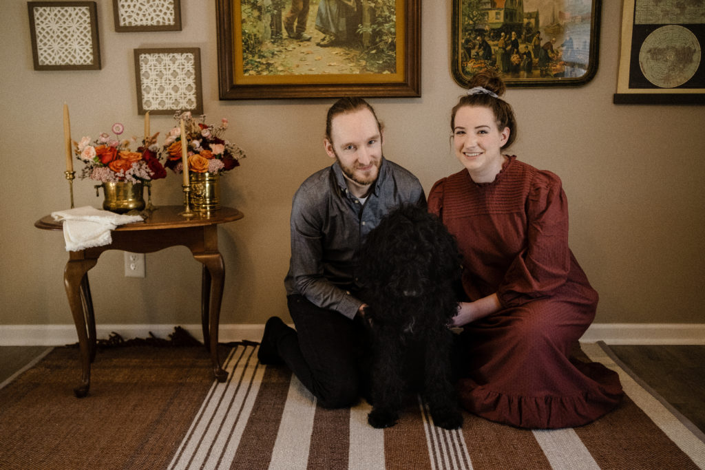 Couple in cottage-core attire posing in front of a gallery wall with their shaggy black dog. Minneapolis couples session.
