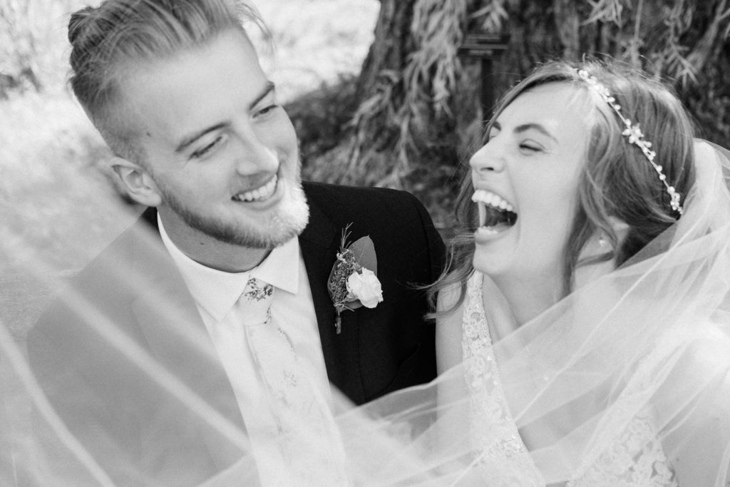 Black and white photo of newlywed couple laughing. They are seated on a bench beneath a willow tree in the Holden Arboretum in Cleveland, Ohio.