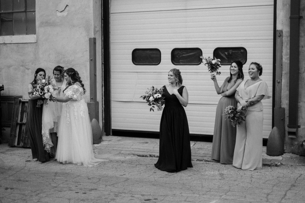 Black and white photo of bride and bridesmaids grouped up while watching and waiting for the groom and groomsmen's portraits to be finished, in an alley downtown Des Moines, Iowa.