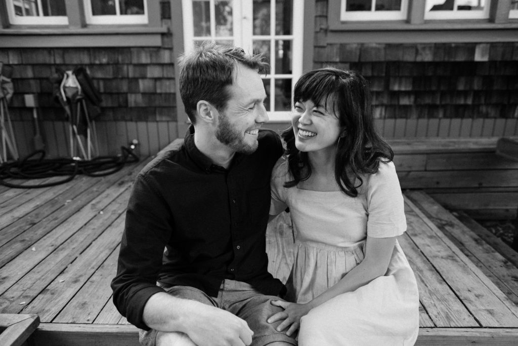 Black and white backyard engagement photo of Craig and Steph sitting side-by-side on their deck smiling at each other. 
