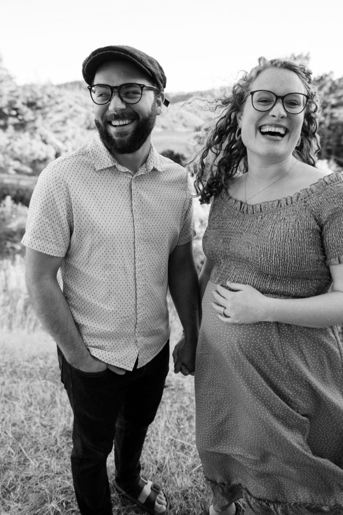 Vertical black and white image of Hannah and Jonah laughing towards the camera, while holding hands. Hannah's hand is resting on her baby bump. California maternity photos.