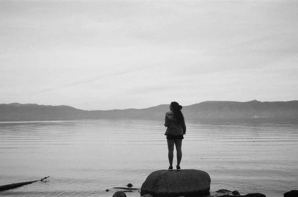 Black and white portrait of Ada standing on a rock facing Lake Tahoe with mountains and fog in the background.