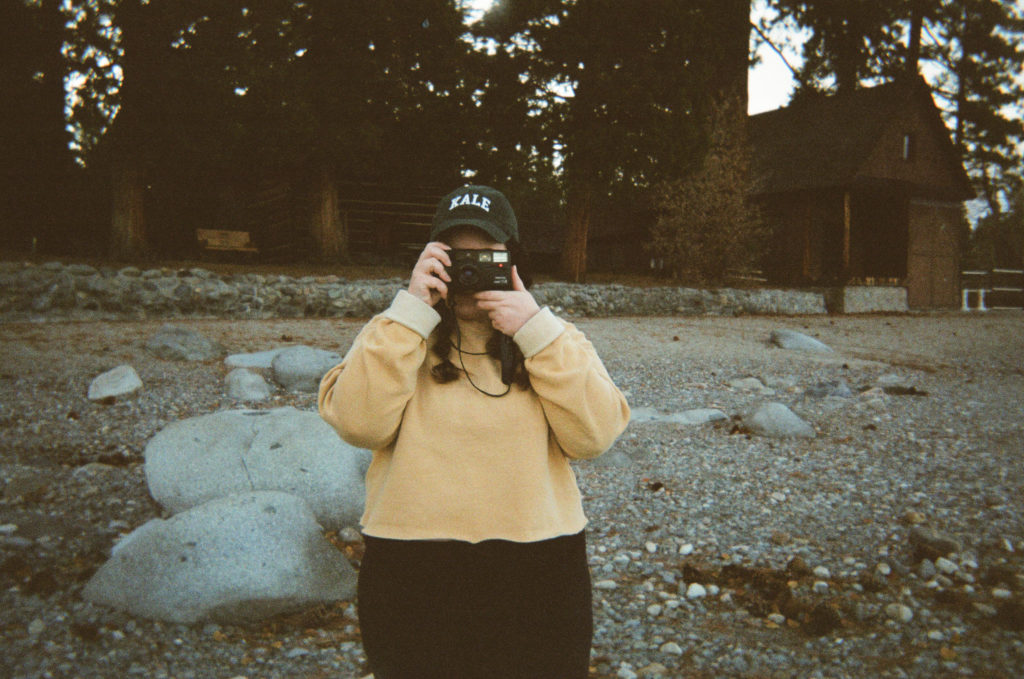 Color photo of Ashley wearing a cropped mustard yellow sweatshirt, braids and a green ball cap holding a film camera.