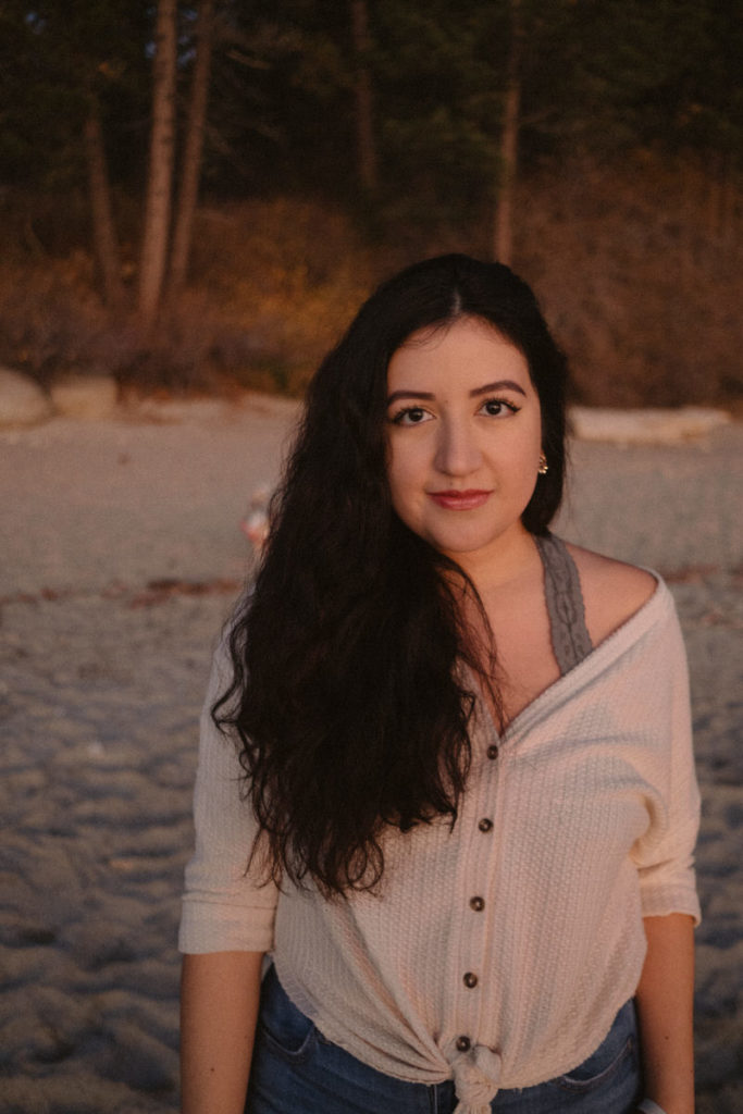 A portrait of Ada, a young Latina woman with long wavy hair, wearing a cream colored top, standing on the beach at sunset on Hidden Beach.