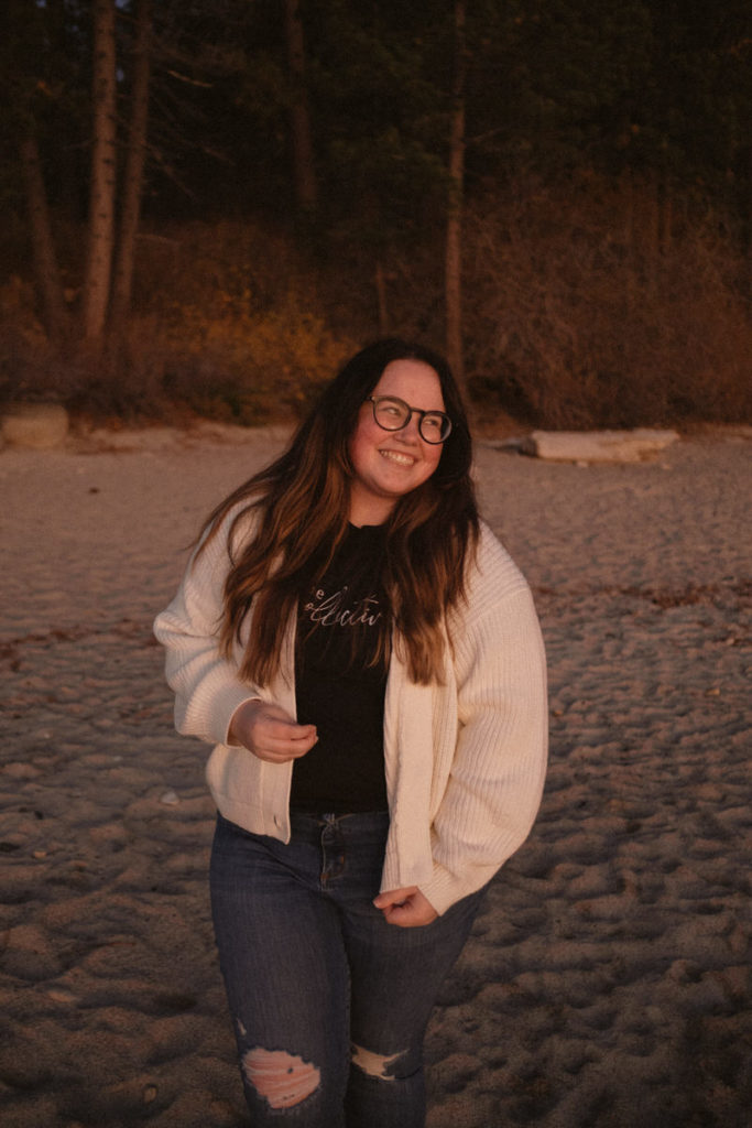 A portrait of Ashley, a white woman with long wavy hair wearing a cream colored cardigan over a black graphic tee, standing on the shore by Hidden Beach at sunset.