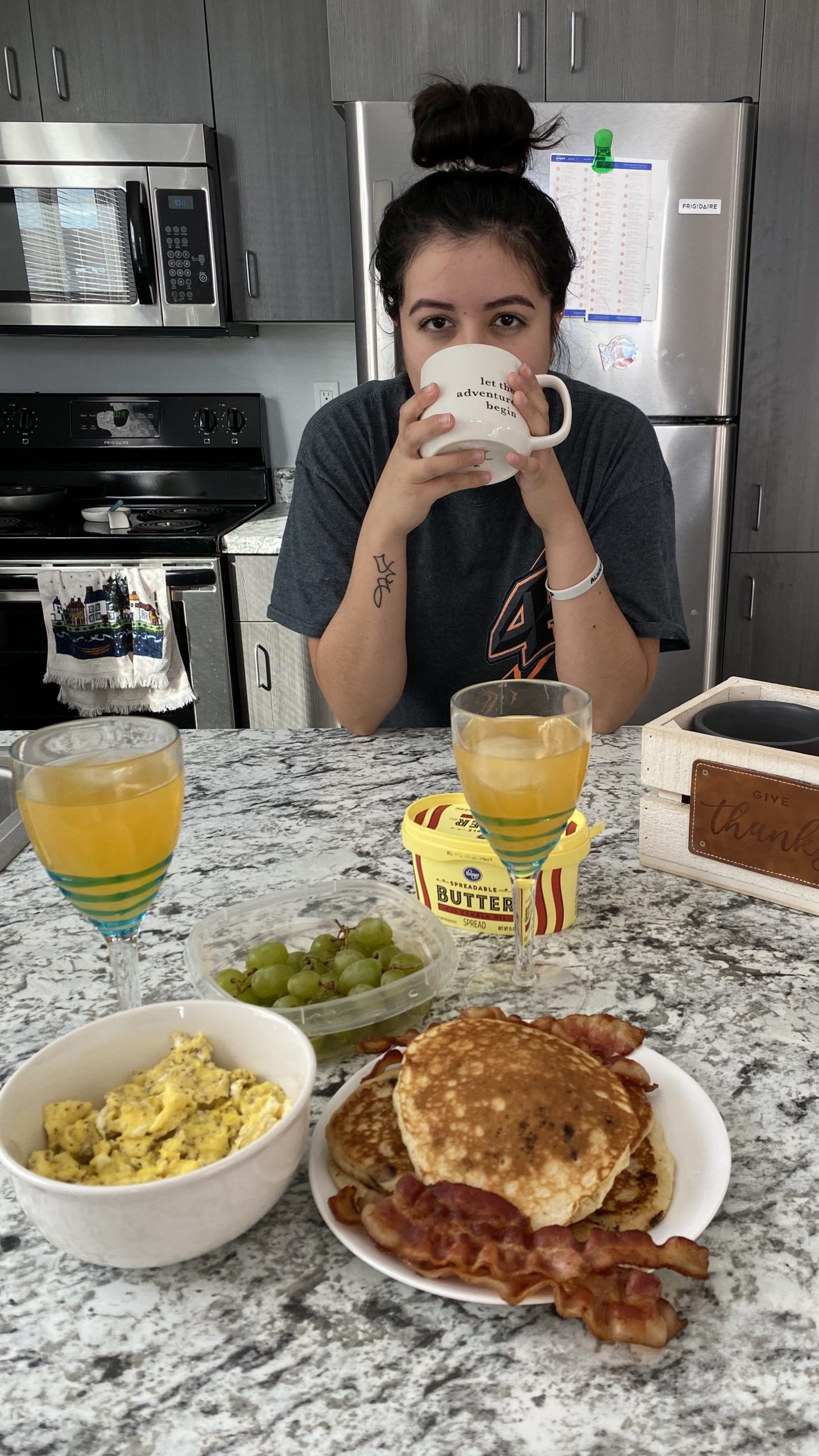 Ada drinking coffee, hair in a bun, and an oversized t-shirt, standing behind the counter where our brunch of eggs, pancakes, and bacon with a side of grapes and juice are laid out. 