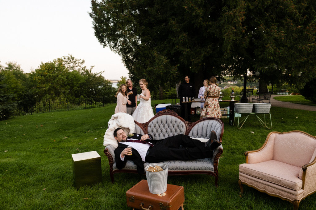 Groom lounging on a vintage sofa at Pioneer Park in Stillwater MN. Colorful photo of an intimate outdoor fall wedding. 
