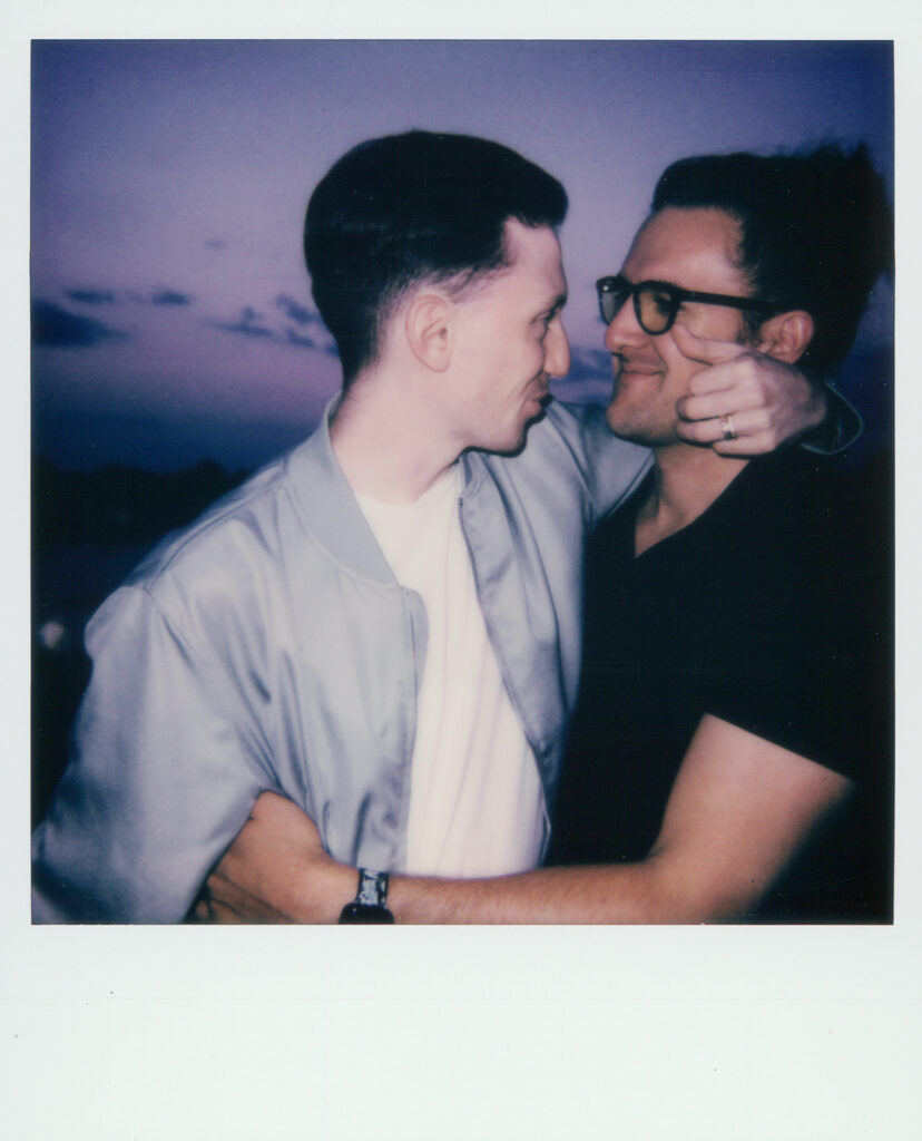 Summer couples photos at Como Park. Ian's arm hangs around Jeremy's neck, cradling his face with his fingers. Jeremy holds onto Ian's waist and they stare sweetly at each other. Photo made on Polaroid at night with flash.