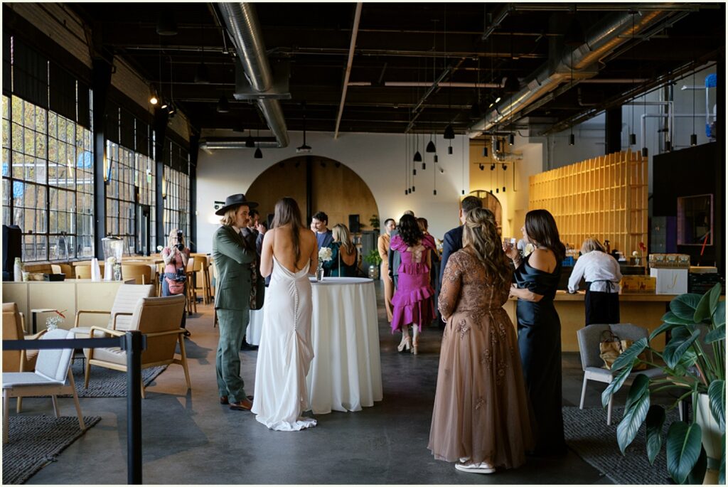 Wedding guests gather in a sunny brewery wedding venue during cocktail hour.